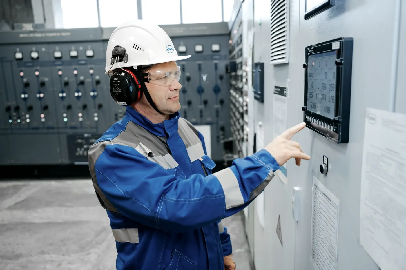 Un opérateur en bleu de travail, casque et protections auditives, pointe l’écran de commande d’une armoire industrielle pour y lire une valeur.