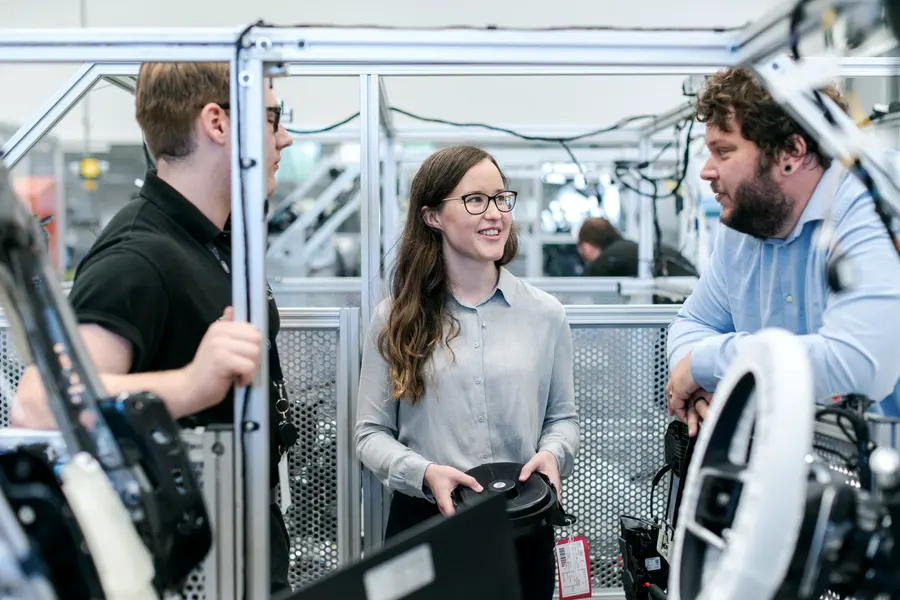 Trois personnes discutent debout devant une machine de production, l’une explique en désignant l’installation.
